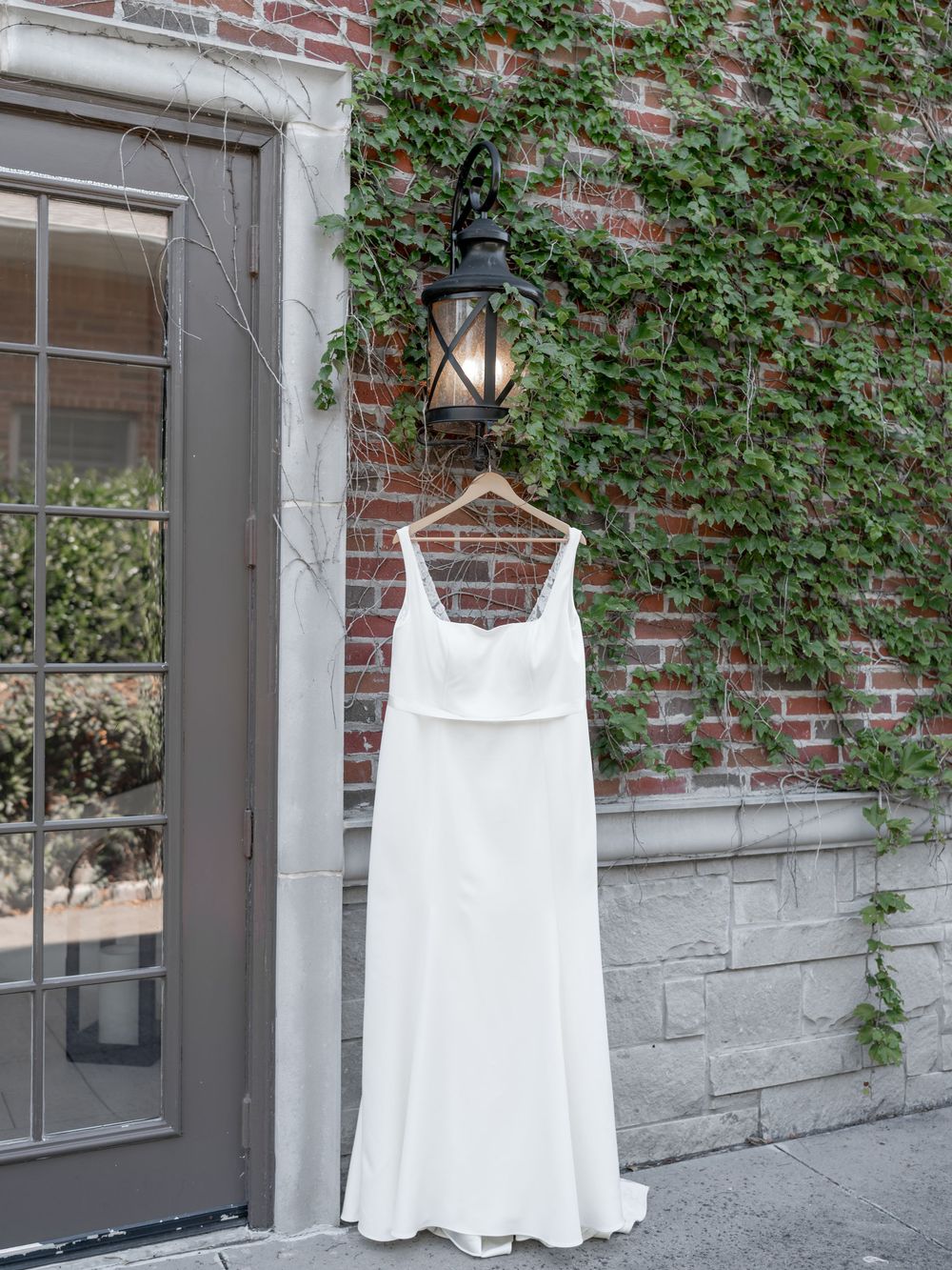 White wedding dress hanging on display against an ivy-covered brick wall with windows. DALLAS WEDDING PHOTOGRAPHER