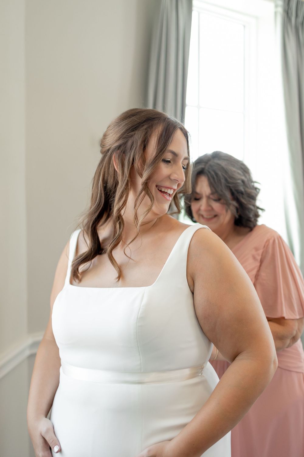 A joyful moment captured during wedding preparations with a woman in a white dress and companion in pink. DALLAS WEDDING PHOTOGRAPHER