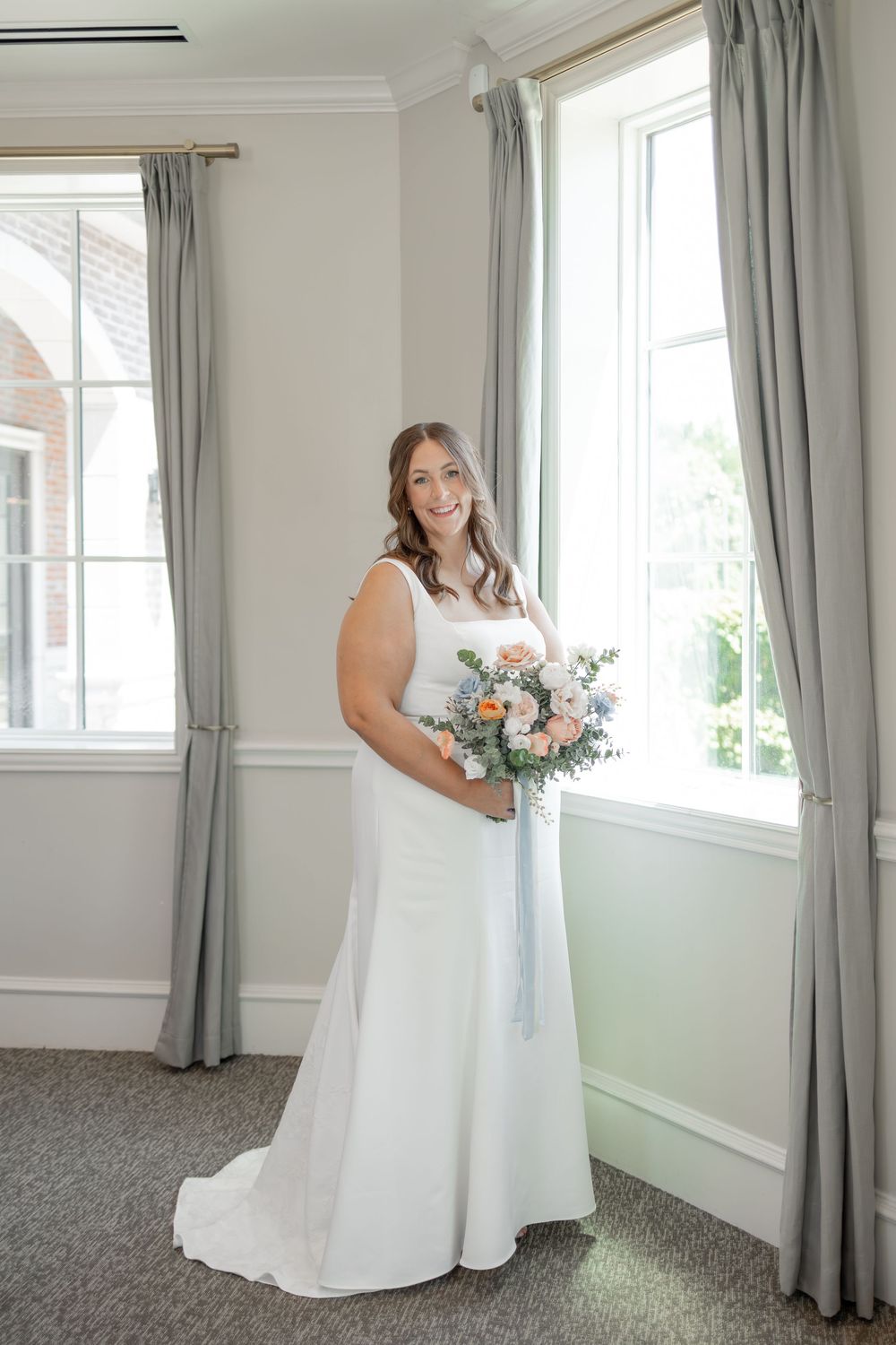 A white wedding dress with a beautiful floral bouquet in a bright room with gray curtains. DALLAS WEDDING PHOTOGRAPHER