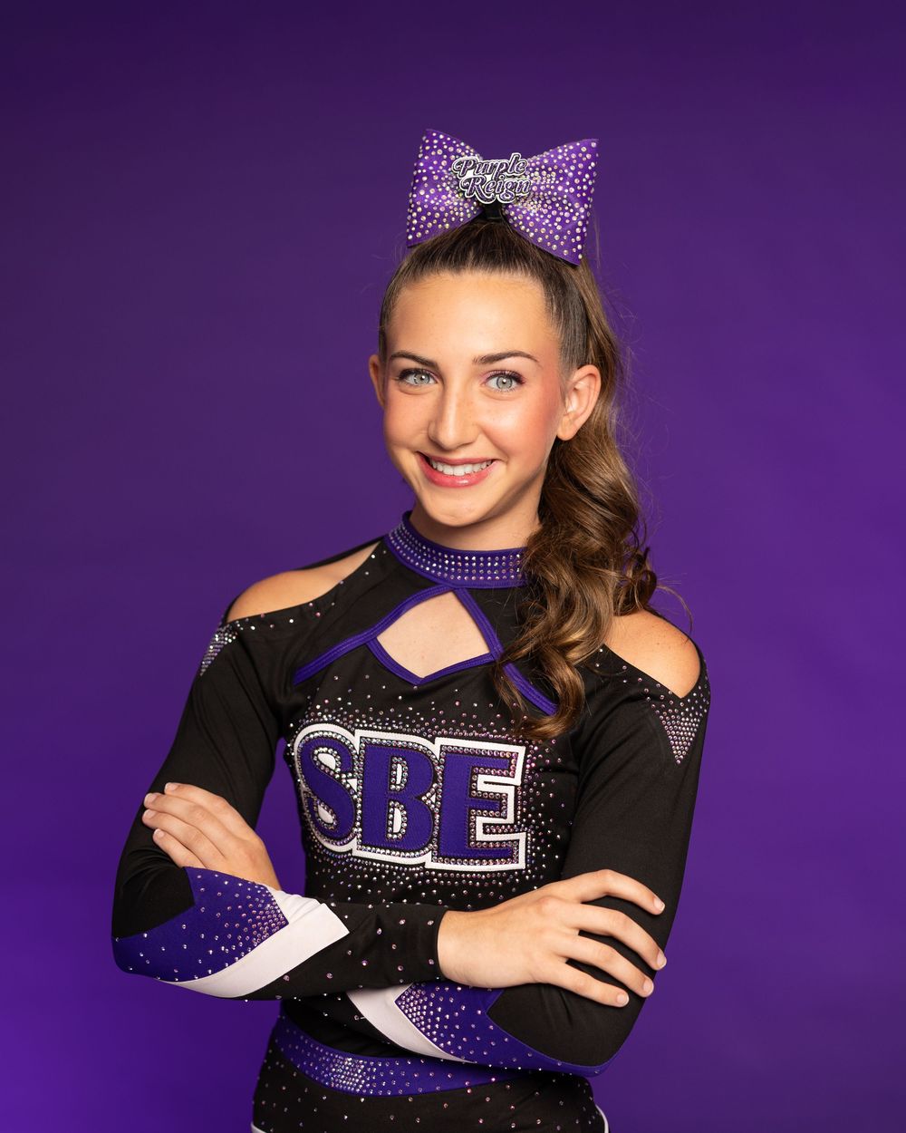 Cheerleader poses in sparkly black and purple uniform with bow against purple backdrop.