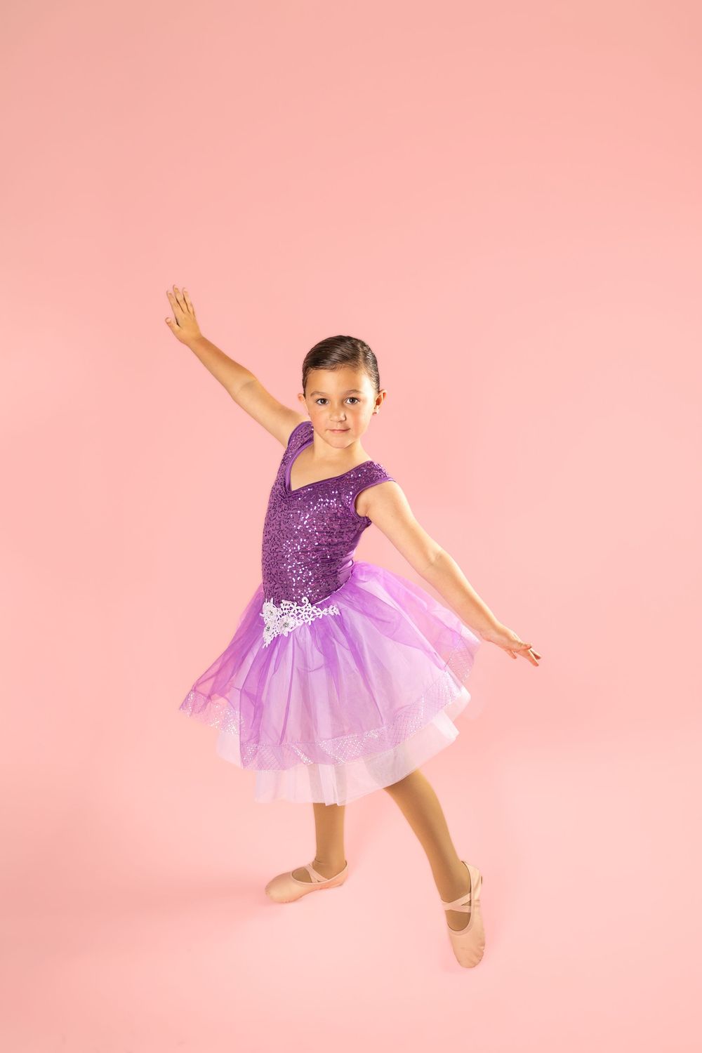 Young dancer in purple tutu performs graceful ballet moves against pink backdrop.