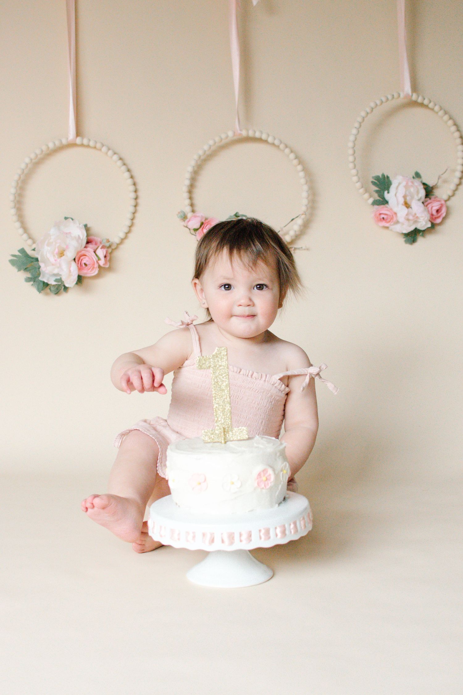 Sweet baby girl in pink romper touching her first birthday cake with floral wreaths in background.