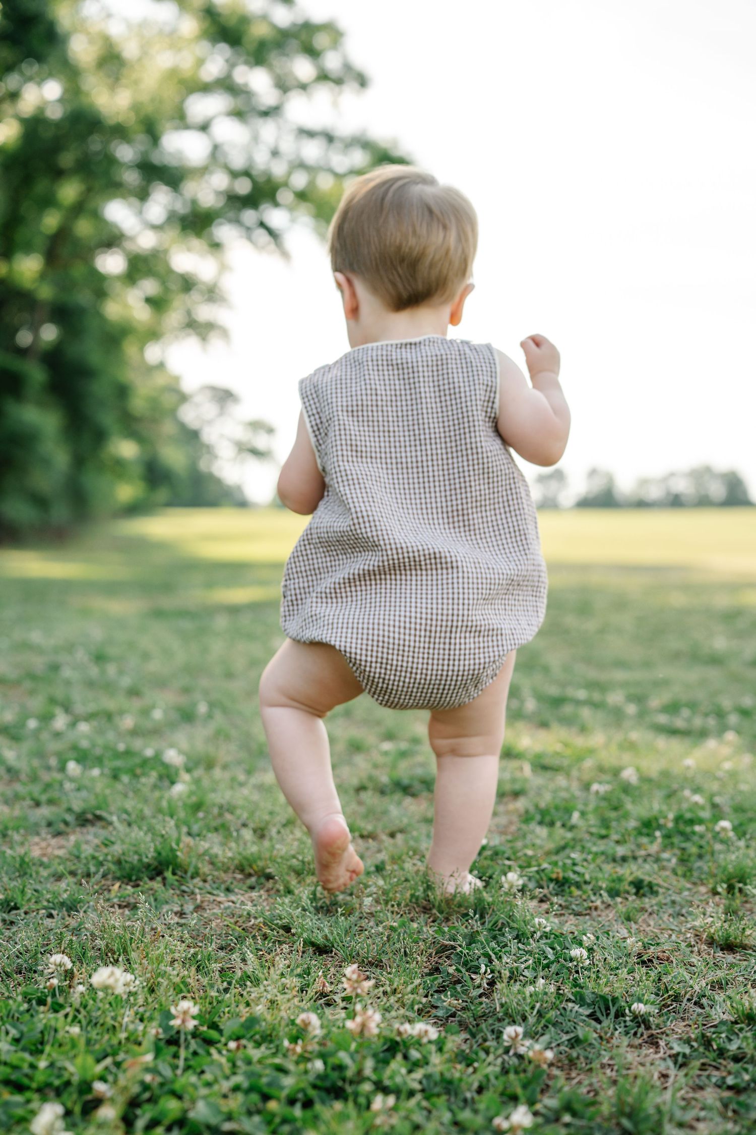 A baby in a plaid romper takes first steps barefoot on green grass in a sunny park.