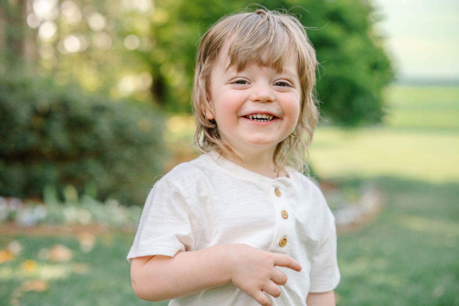 Happy toddler with blonde hair smiling outdoors in a white t-shirt on a sunny day in a green park.