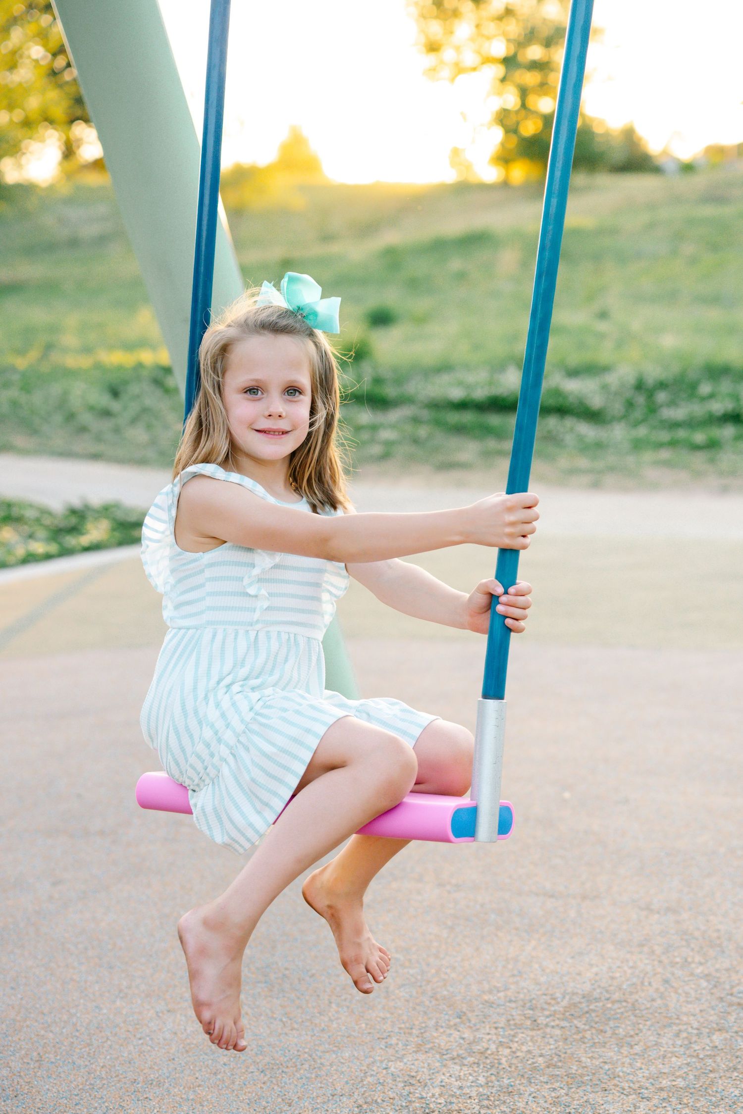 A smiling young girl in a striped dress sits barefoot on a colorful playground swing at sunset.