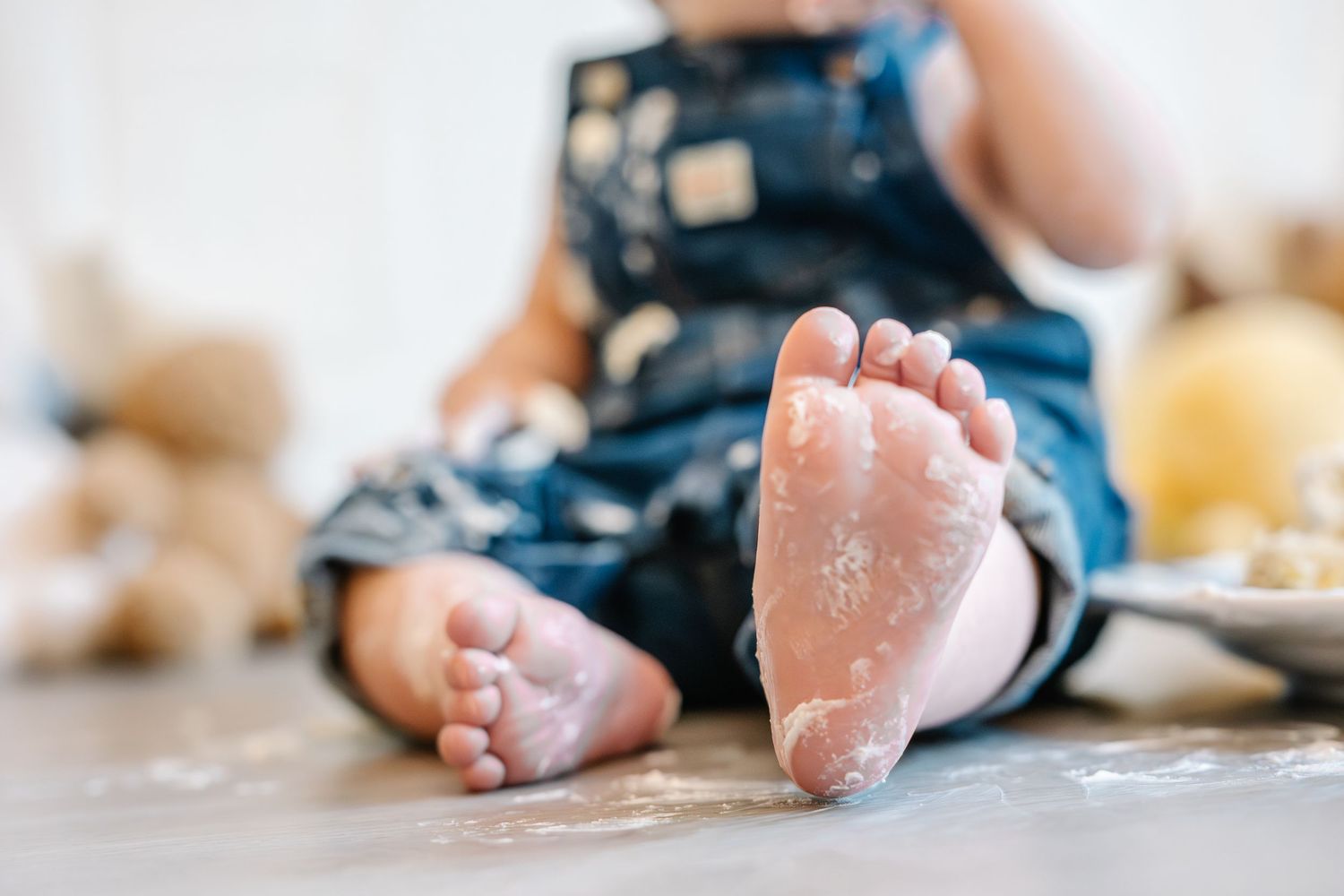 Close-up of a baby's flour-covered feet sitting on the floor in a denim outfit with toys in the background.
