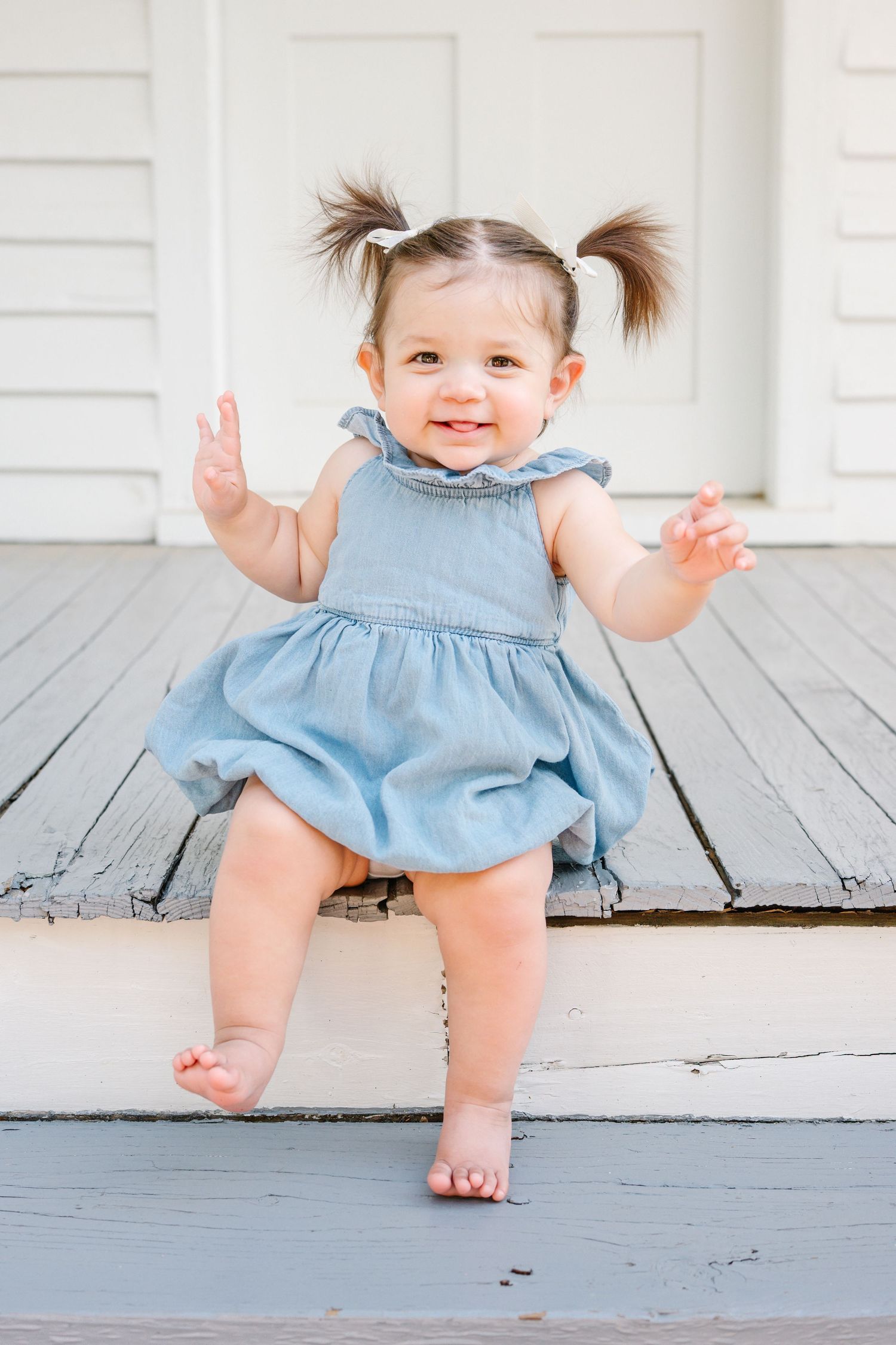 Smiling baby girl in blue denim romper with pigtails sitting on wooden porch steps, hands raised playfully.