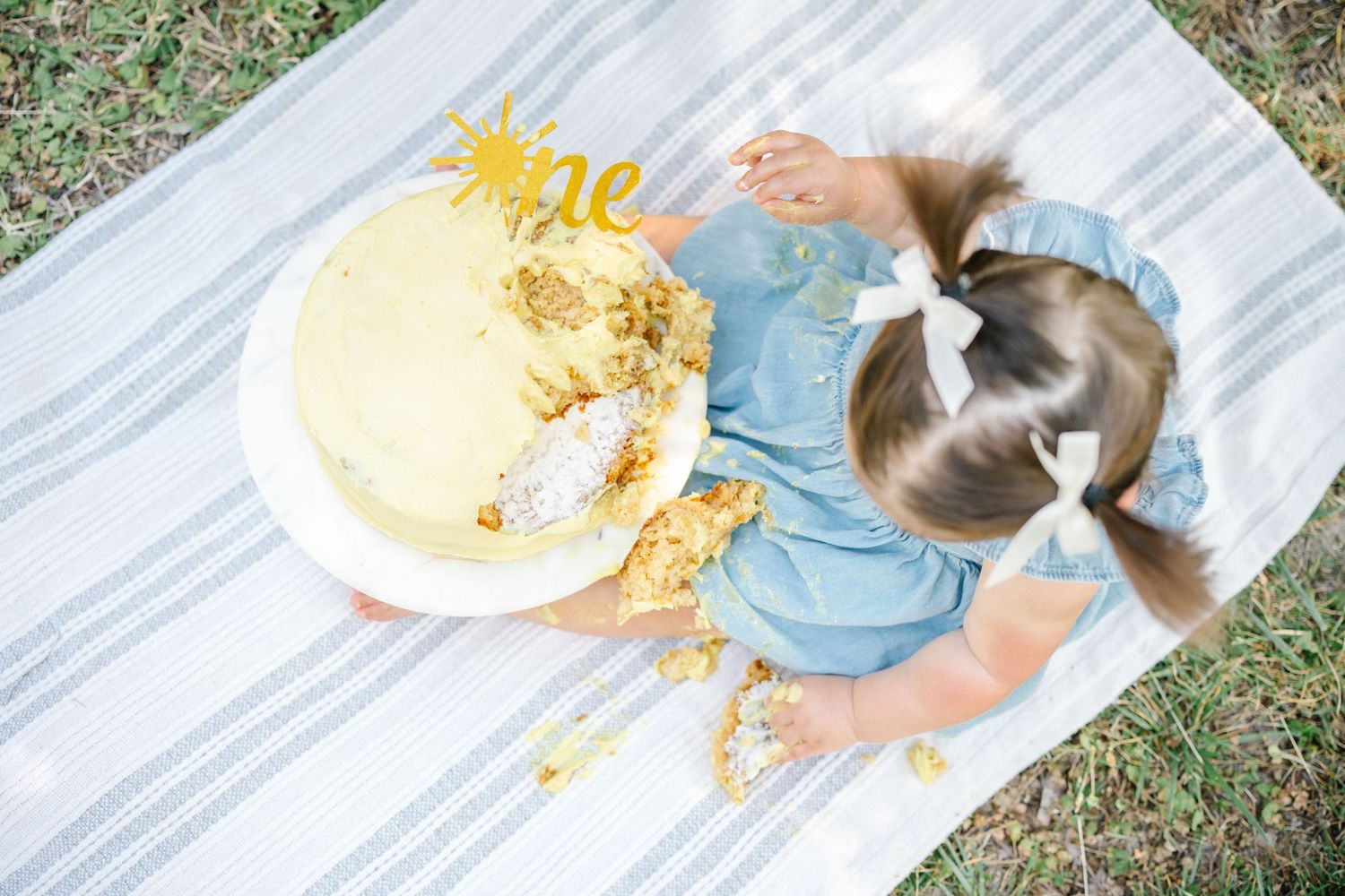 Aerial view of a toddler girl in a blue dress with pigtails smashing her first birthday cake outdoors.