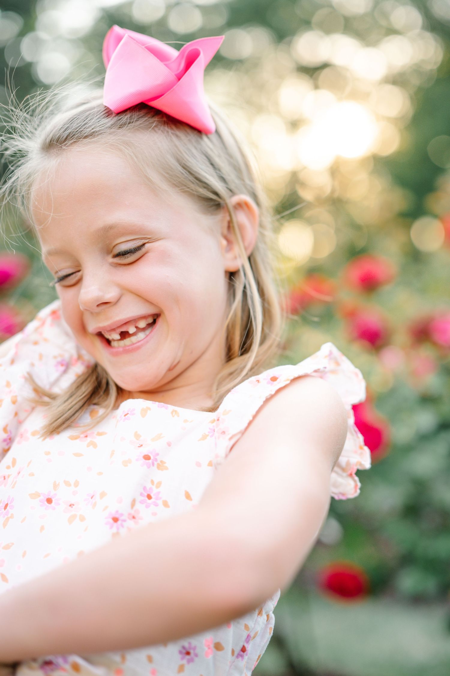 A joyful young girl with a pink bow laughs outdoors, wearing a floral dress near blooming red roses.