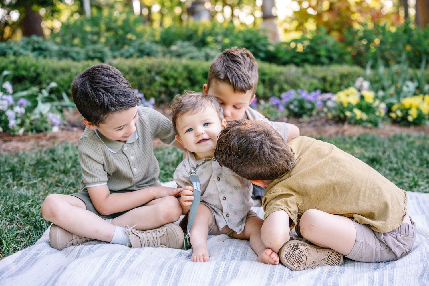 Four young brothers sitting on a blanket outdoors, sharing a sweet moment together surrounded by a lush garden.