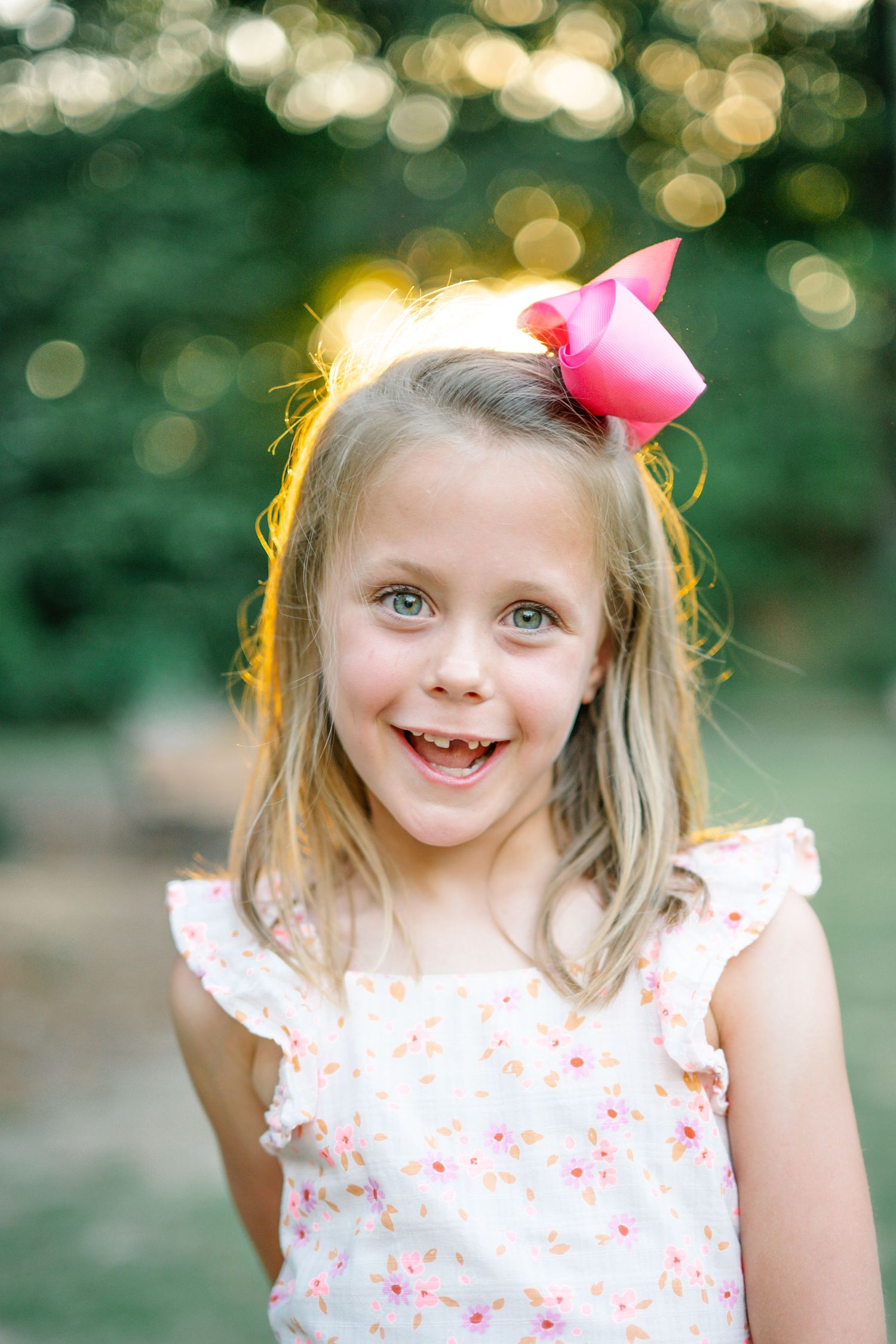 Smiling young girl with blonde hair wearing a pink bow and floral dress, posing outdoors with bokeh background.