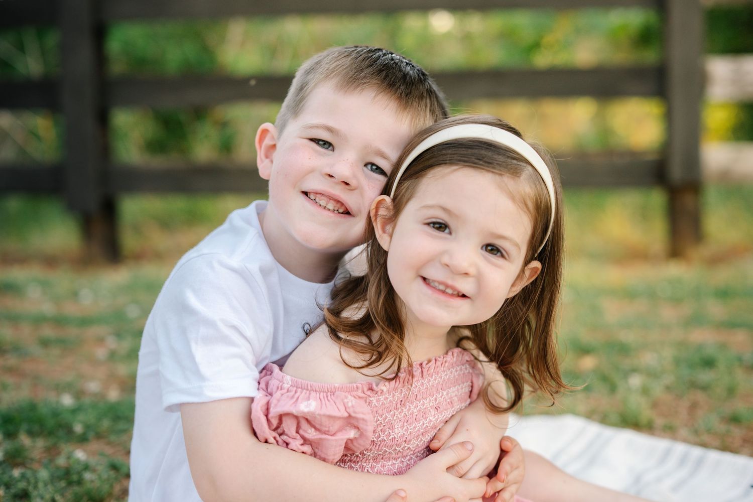 Two smiling children, a boy and girl, hugging outdoors near a wooden fence on a sunny day.