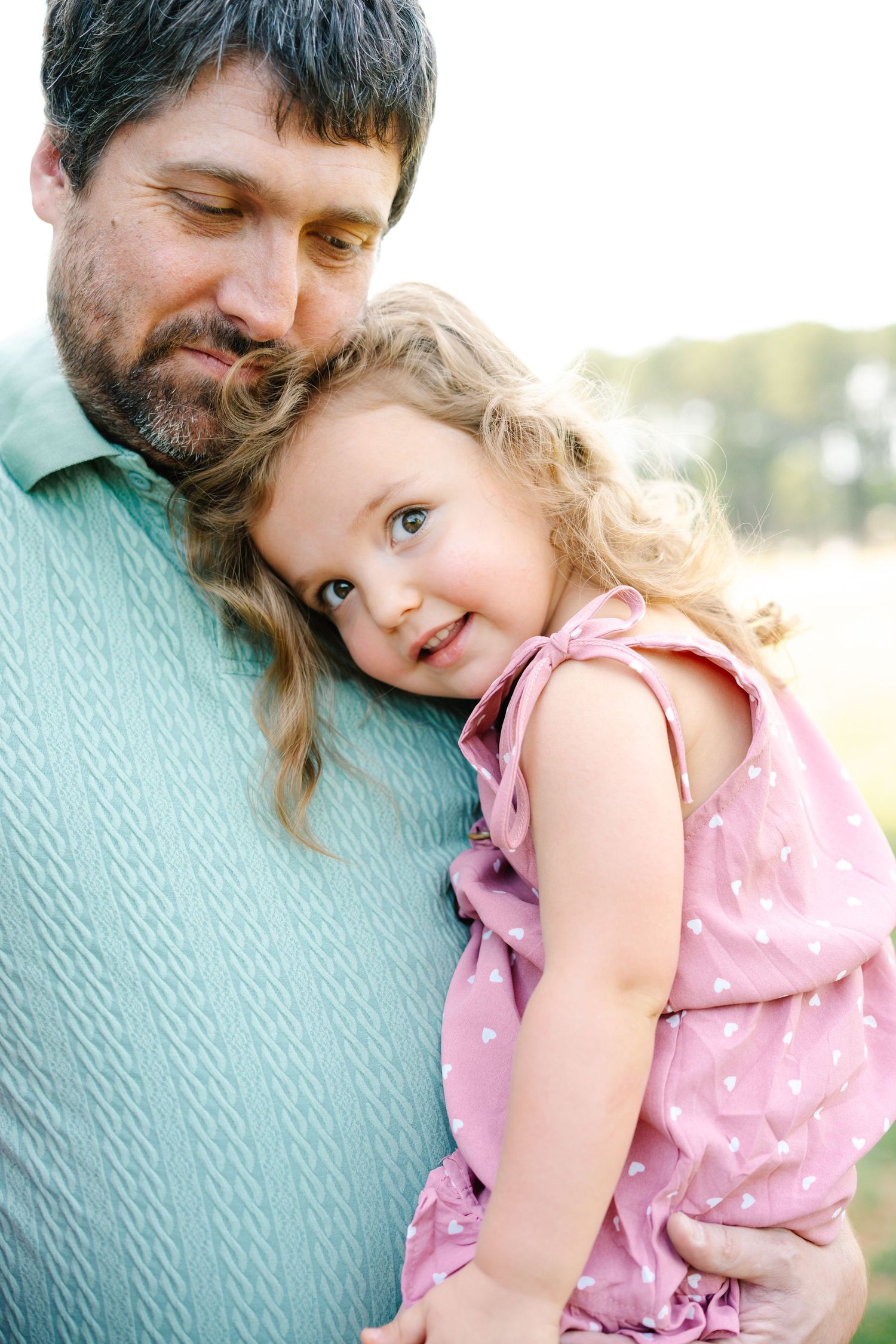 A bearded father tenderly holds his smiling curly-haired little girl in a pink polka dot dress outdoors.