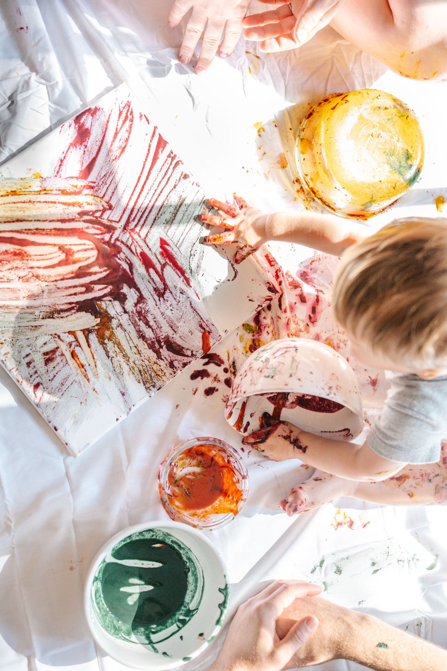Child finger painting with red, orange, yellow, and green paints on white canvas, creating colorful abstract art.