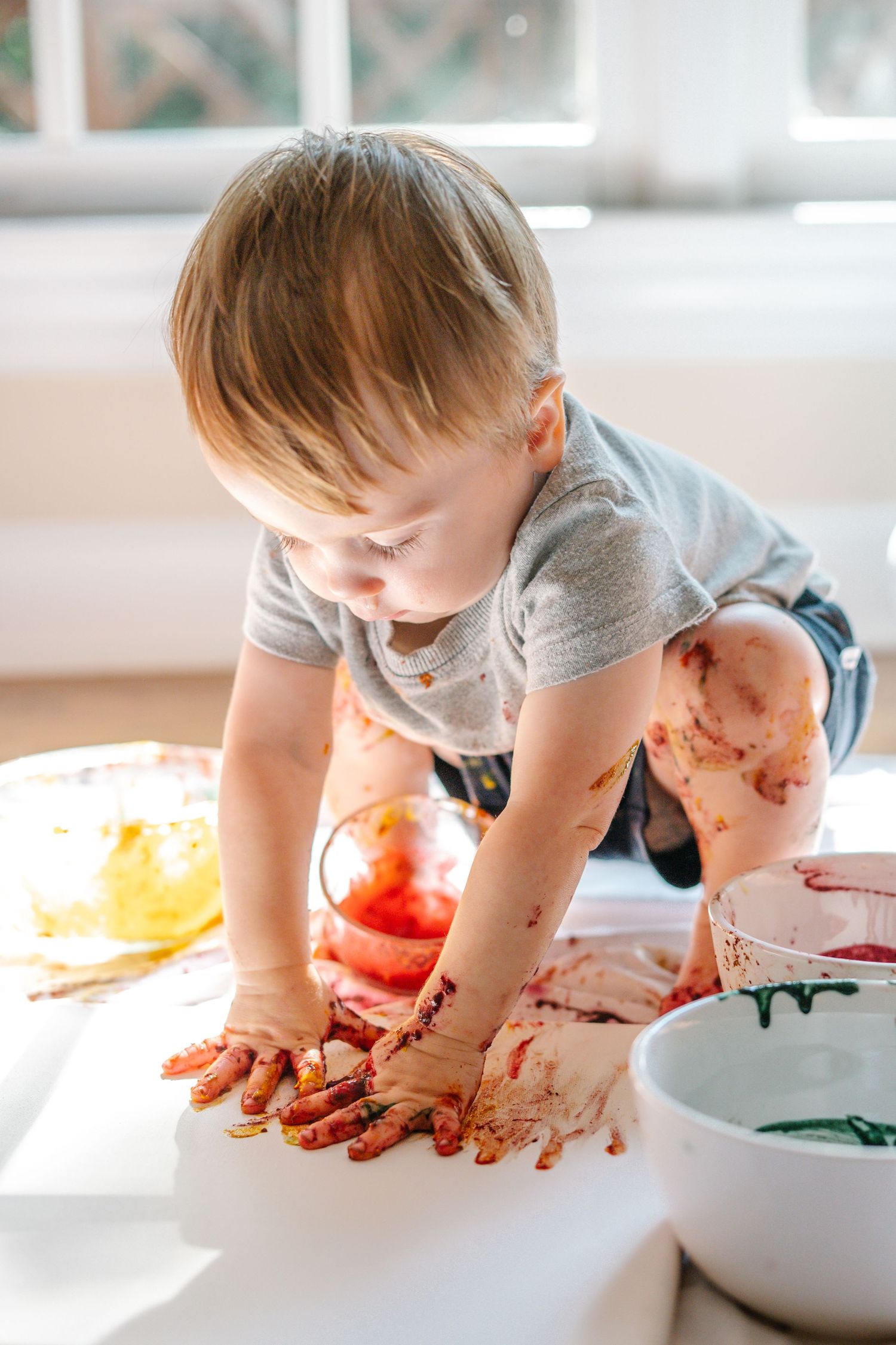 Toddler with paint-covered hands doing finger painting with colorful red, yellow, and green paints on white paper.