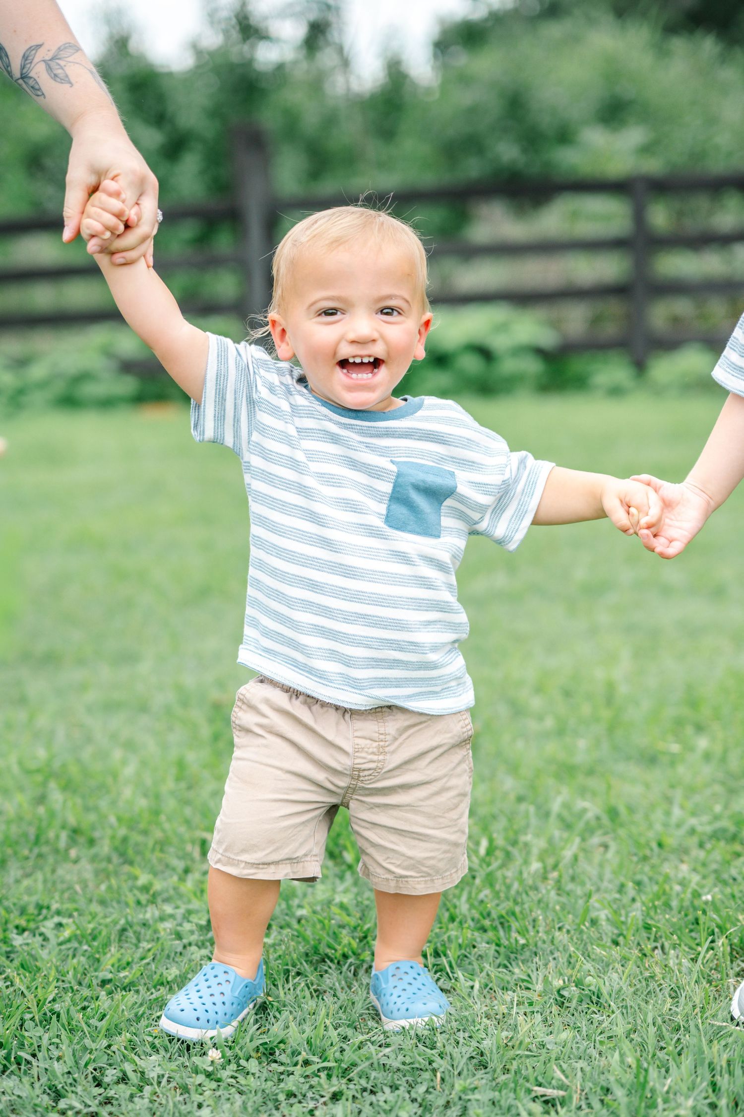 Happy toddler boy in striped shirt and khaki shorts holds hands with adults while standing on green grass outdoors.