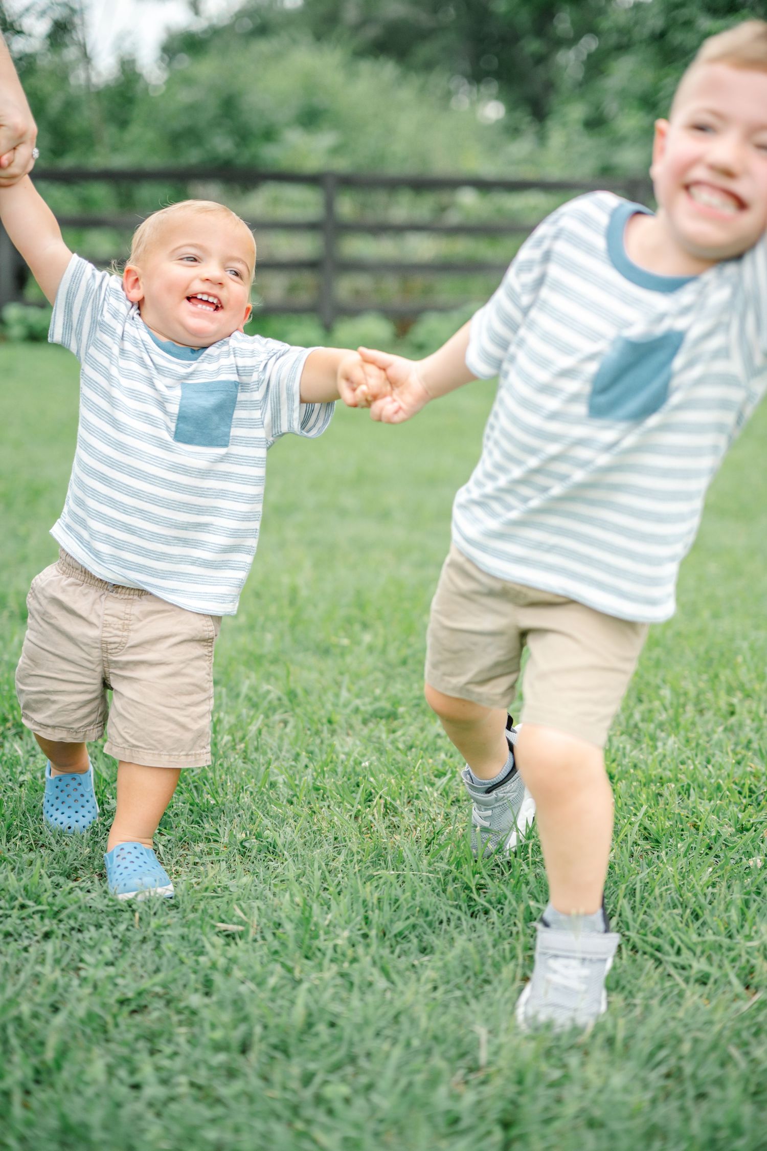 Two young brothers in matching striped shirts and khaki shorts hold hands while playing on green grass outdoors.