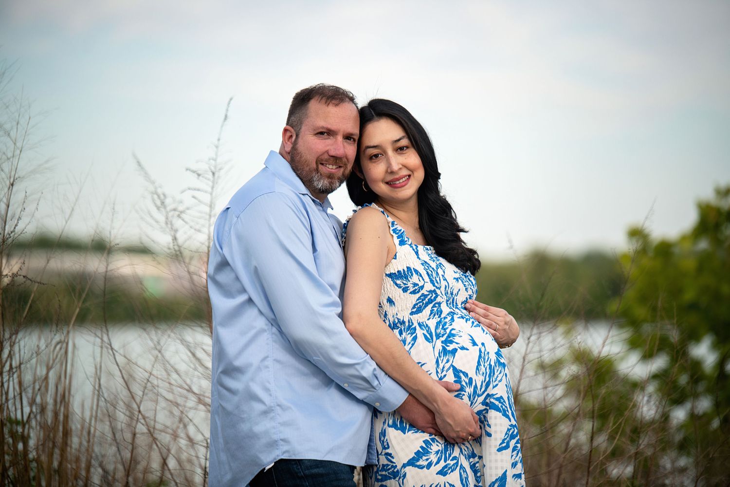 Expectant couple embraces outdoors near waterfront during sunset.