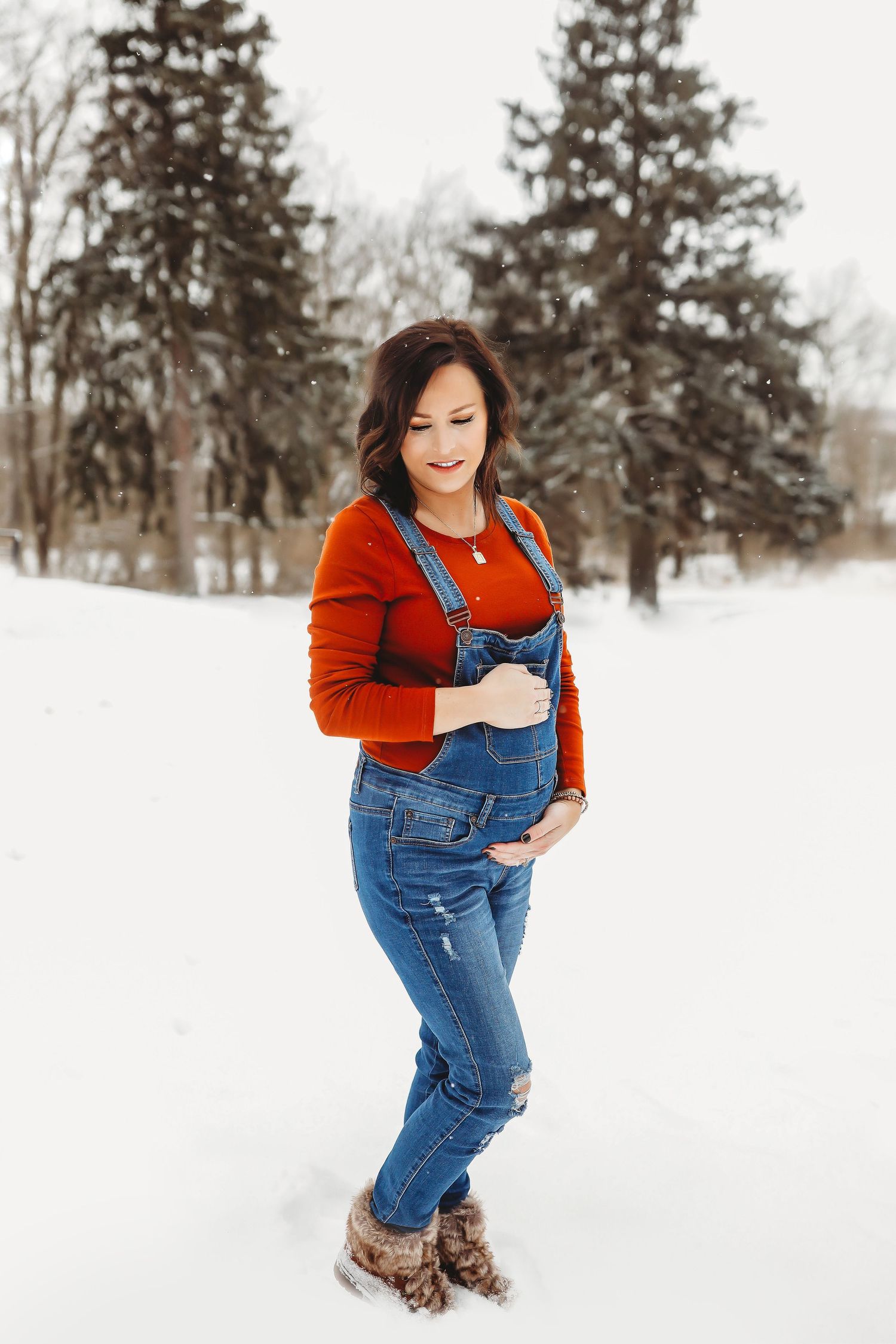 Pregnant woman in denim overalls and red sweater posing in snowy winter park with evergreen trees.