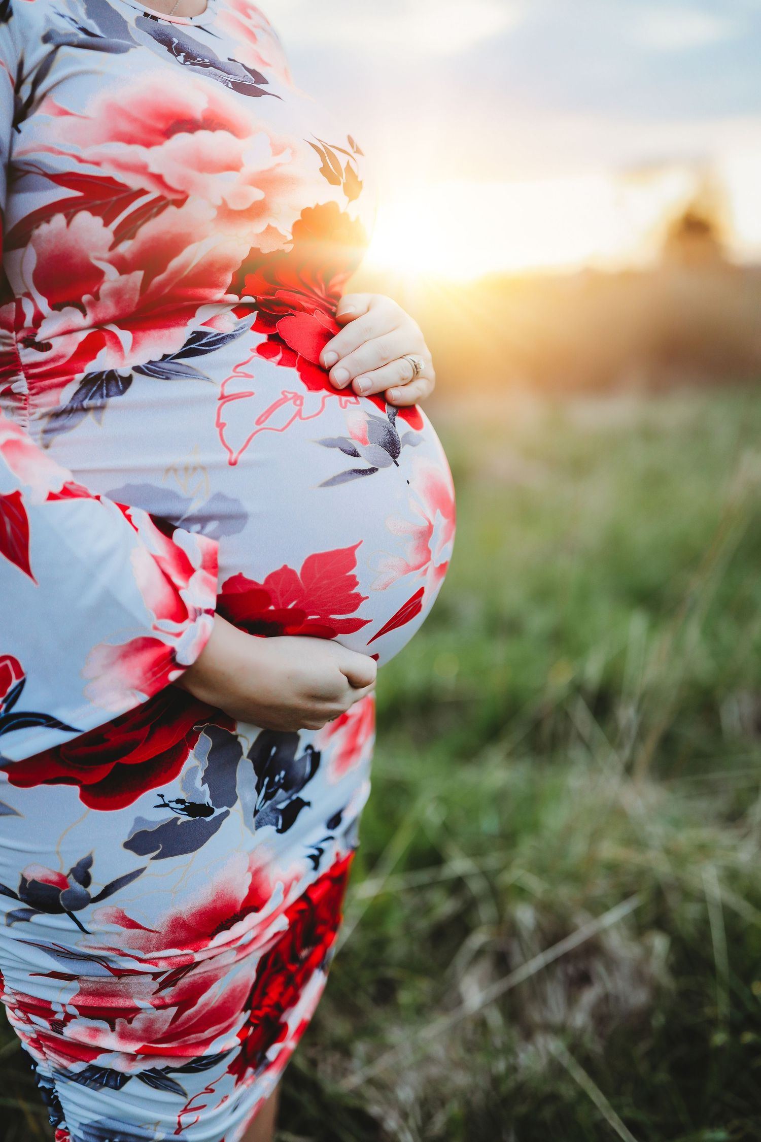 Pregnant woman in floral dress cradling baby bump outdoors at golden sunset in a green field.