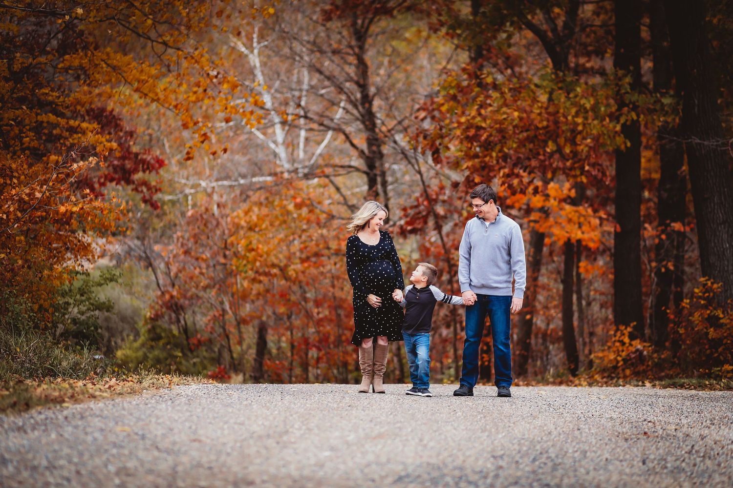 Family of three posing on a gravel path surrounded by vibrant autumn foliage in shades of red and orange.