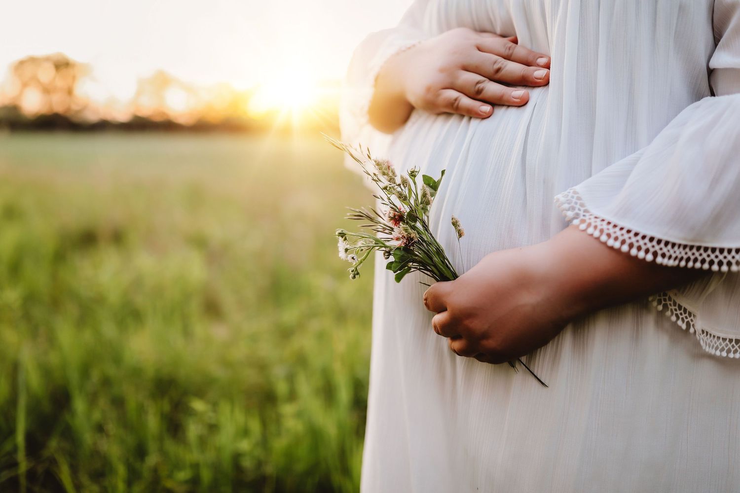Pregnant woman in white dress holding wildflowers in a sunlit green field at golden hour.