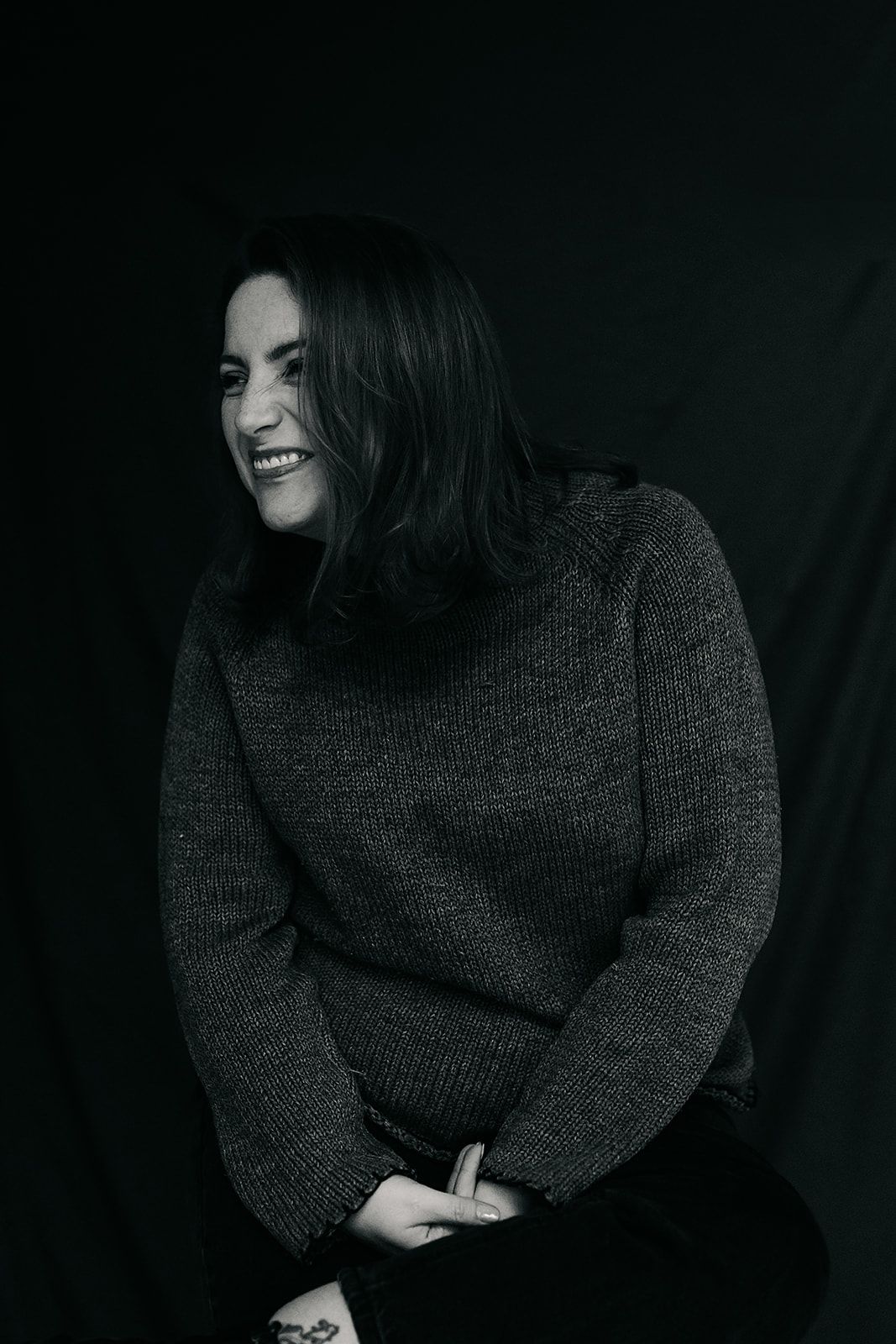 A person wearing a dark knit sweater sits in moody lighting while laughing and looking down in a series of artistic shots. headshot author