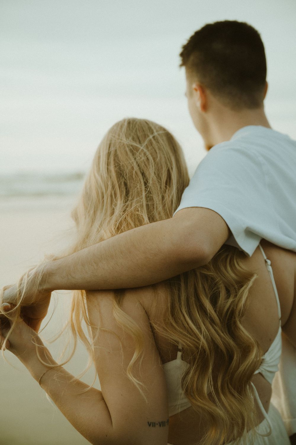 Side view of couple embracing on beach with flowing blonde hair catching the sunset light.