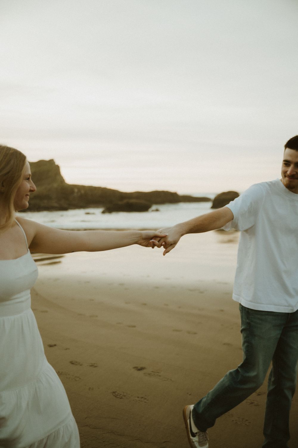Couple holds hands while walking on beach with arms outstretched at sunset.