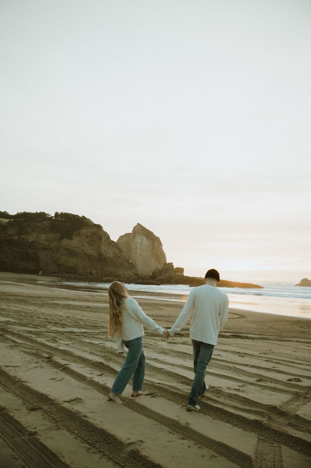 Couple walks hand in hand along a textured beach with coastal cliffs in the distance.