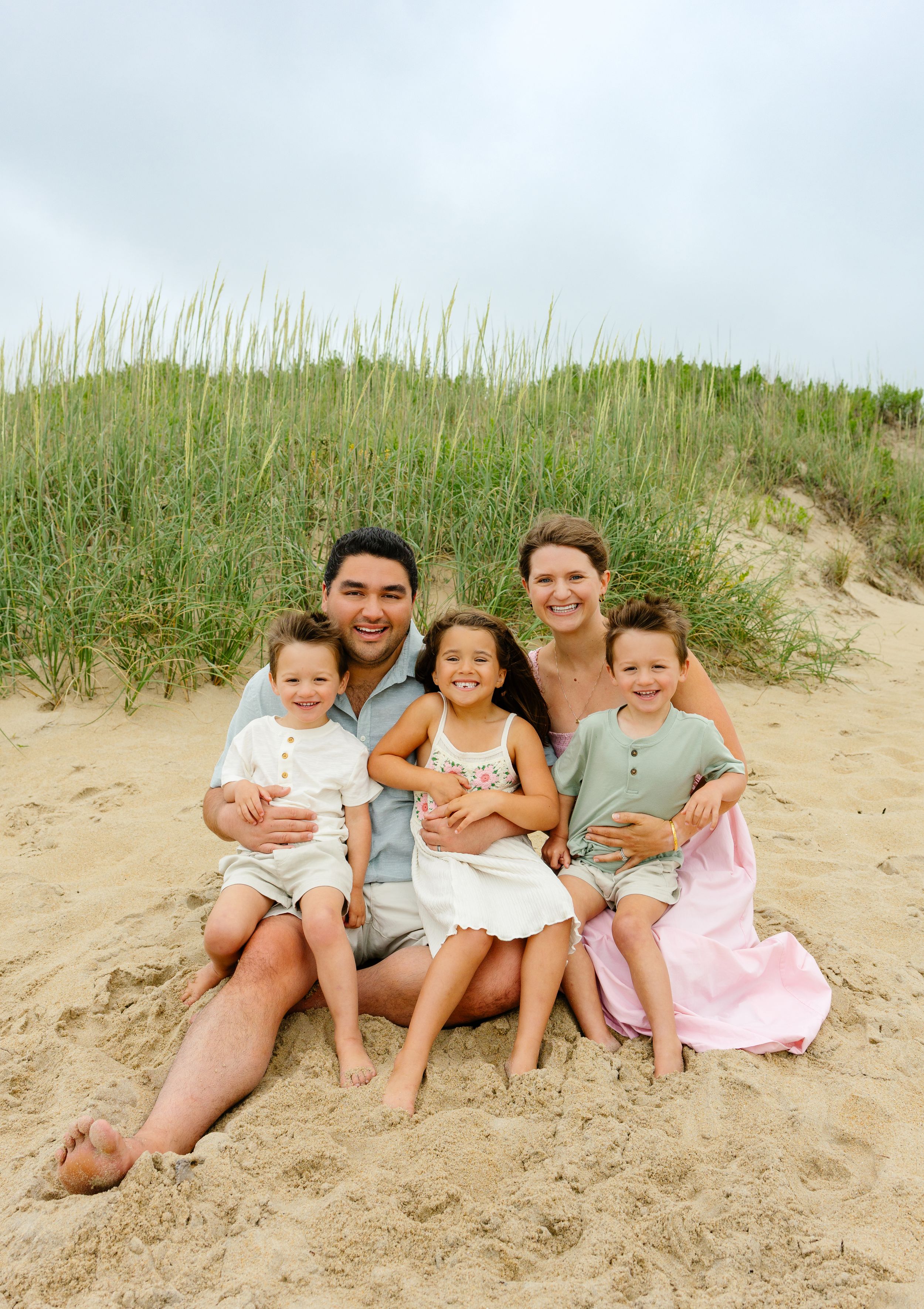 Happy family of five sitting on sandy beach dunes, smiling together on a cloudy day.
