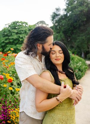 Couple embracing in a garden with colorful flowers, man kissing woman's cheek as she smiles with eyes closed.