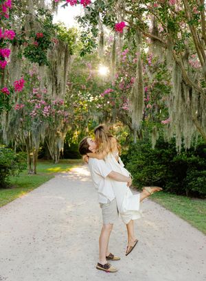 Couple sharing a romantic lift and kiss on a sunlit garden path lined with Spanish moss and pink flowering trees.