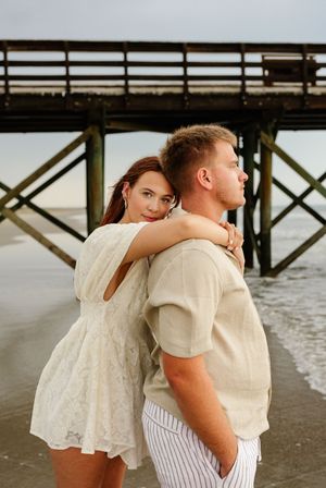 Couple embracing on beach near pier, woman in white lace dress looks back, man in beige shirt and striped pants.