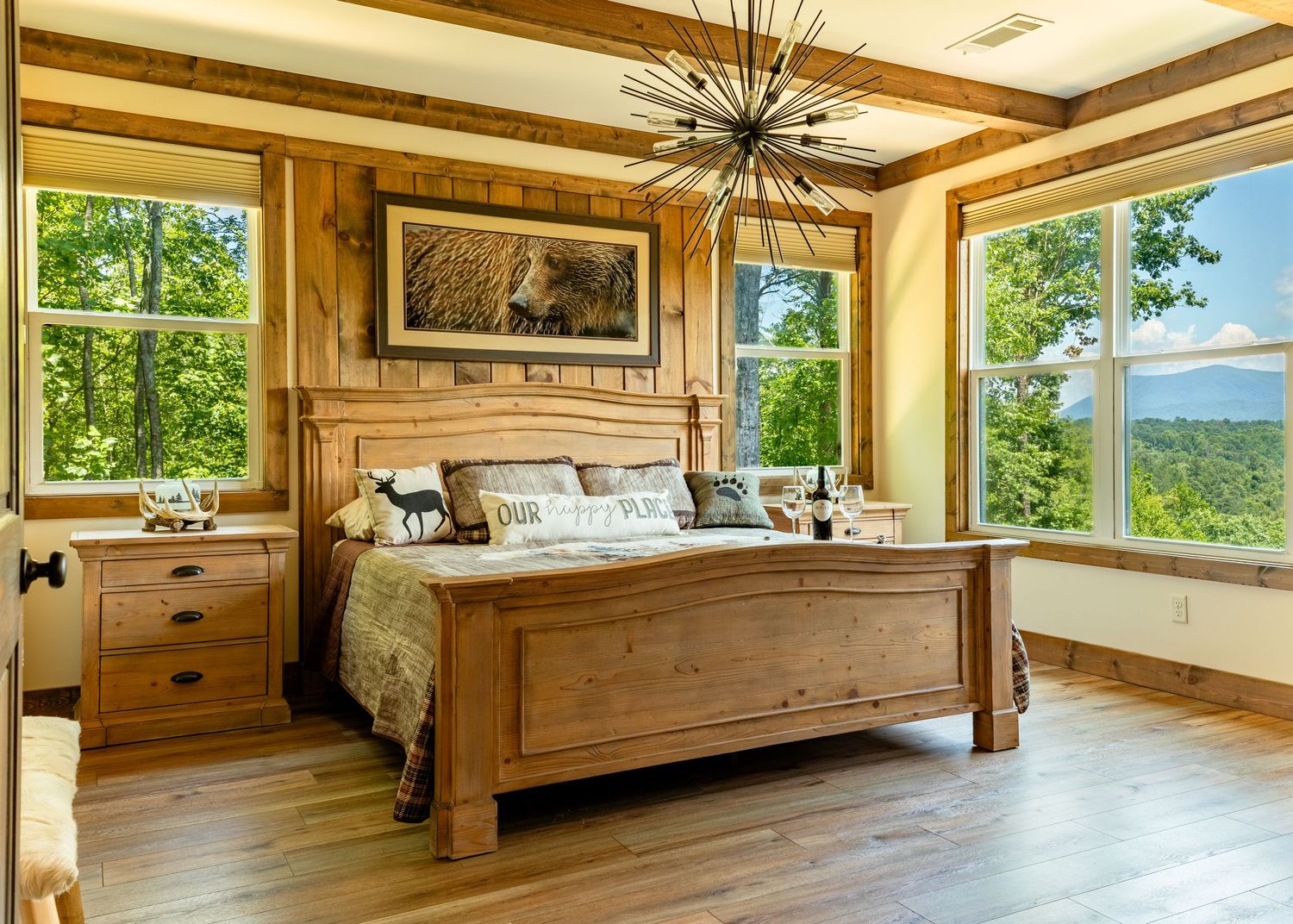 A bedroom with wood accents and large windows showing a vista of trees and blue mountain peaks in the background.