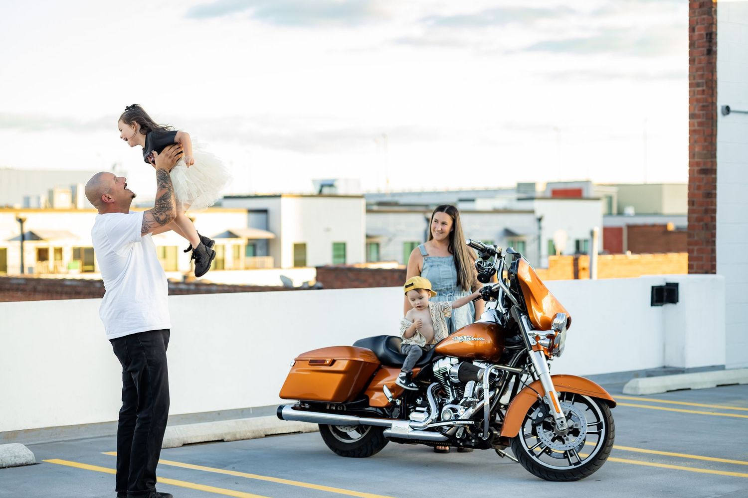 A motorcycle and couple stand on a rooftop parking garage against an urban skyline at sunset.