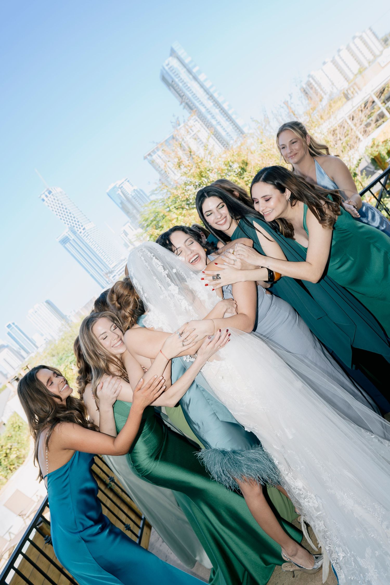 Wedding party in emerald green dresses and a bride in white posing on a sunny rooftop with city skyline during a destination wedding.