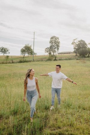 Couple playfully interacting while walking in tall grass meadow on overcast day.