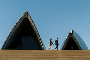 Two people walk along the top of the iconic Sydney Opera House steps against a bright blue sky.