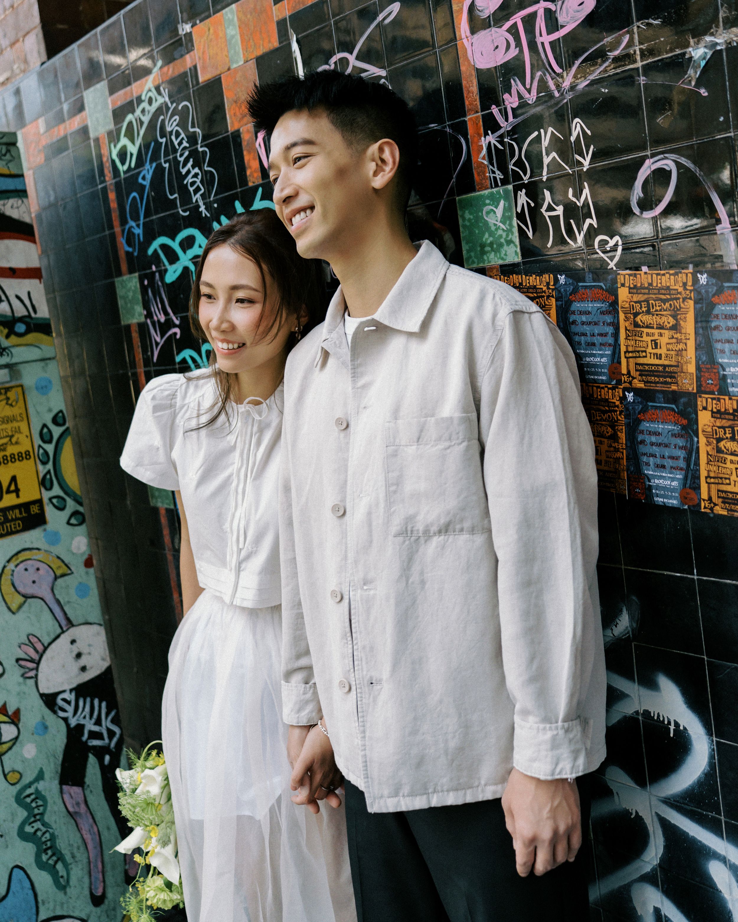 A couple in white outfits share joyful moments against a graffiti wall background.