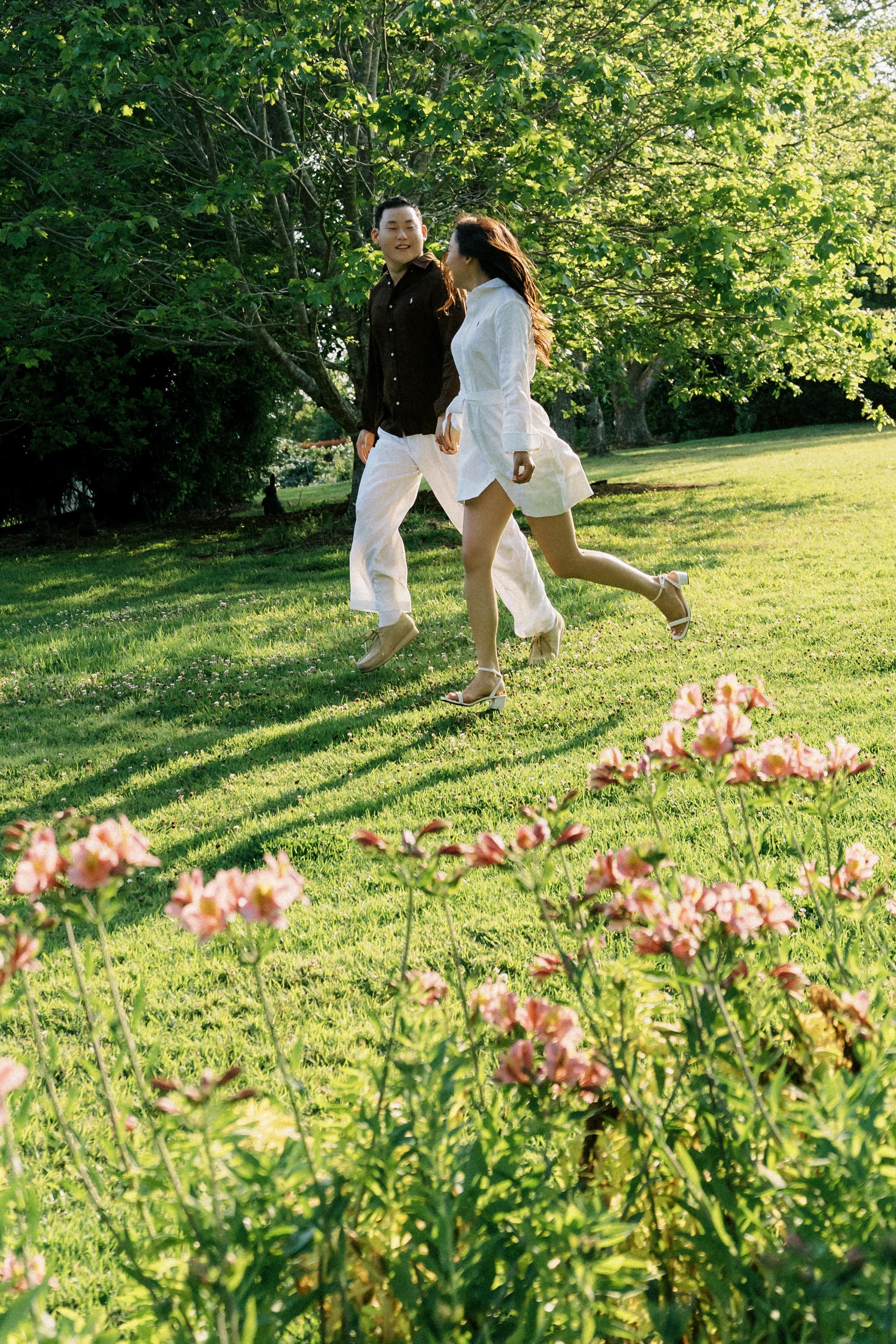 People in white clothing playfully tumbling on a grassy hillside with pink flowers blooming in the foreground.