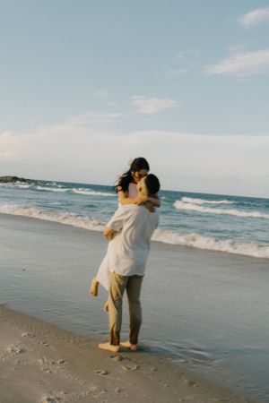 A couple embraces on the beach at sunset while waves crash against the shoreline in a romantic seaside moment.