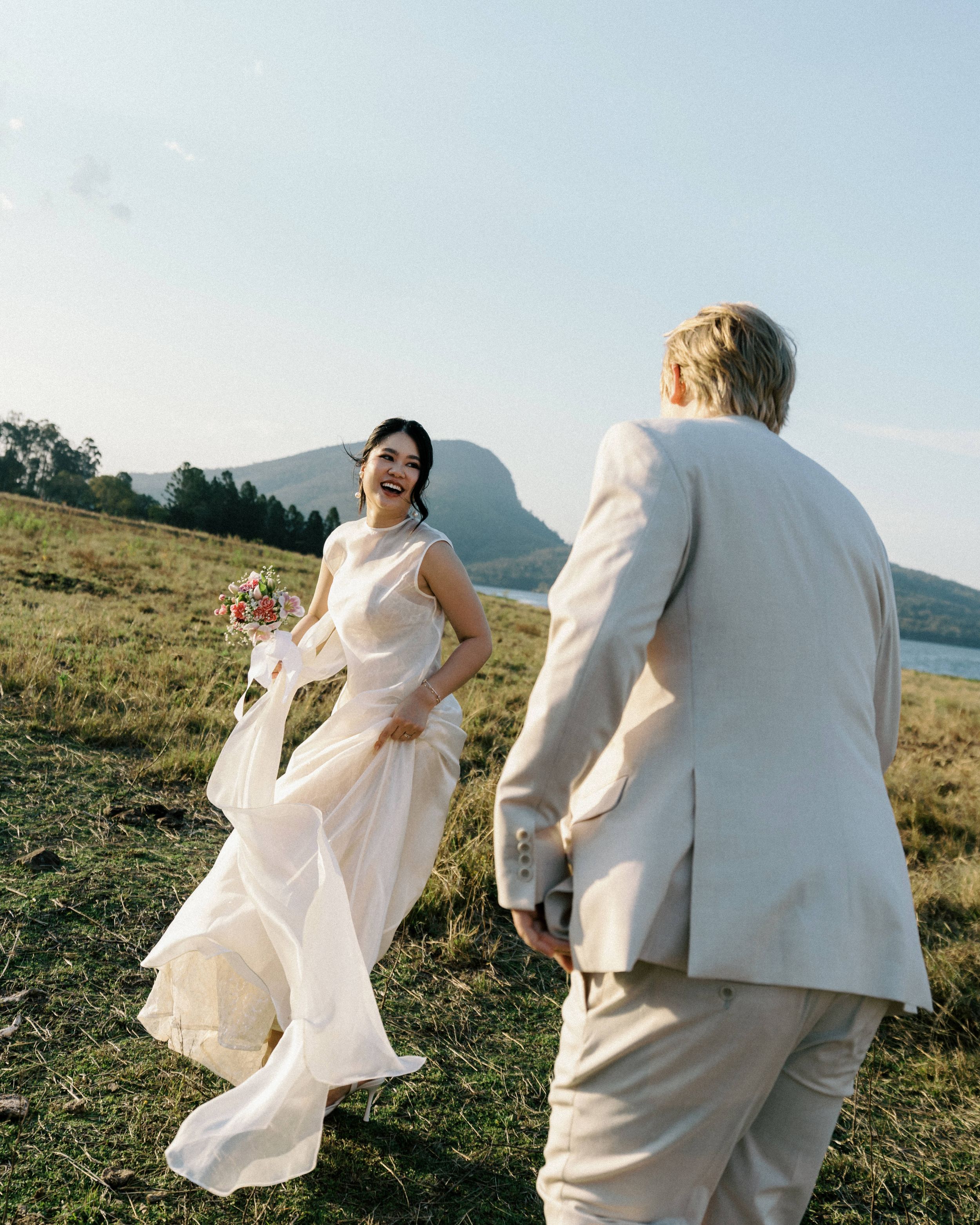 A couple in white outfits dances together on a grassy hilltop overlooking a scenic landscape at sunset.