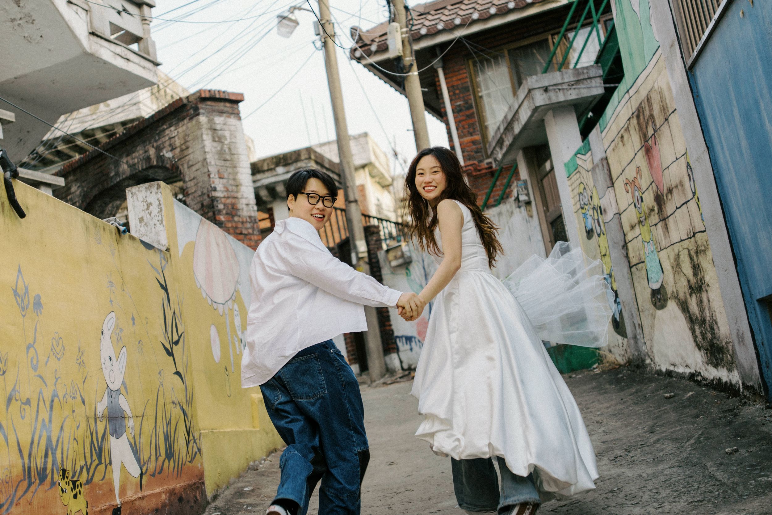 Couple in wedding attire holding hands in a colorful Korean alley, bride in white dress, groom in jeans and white shirt.