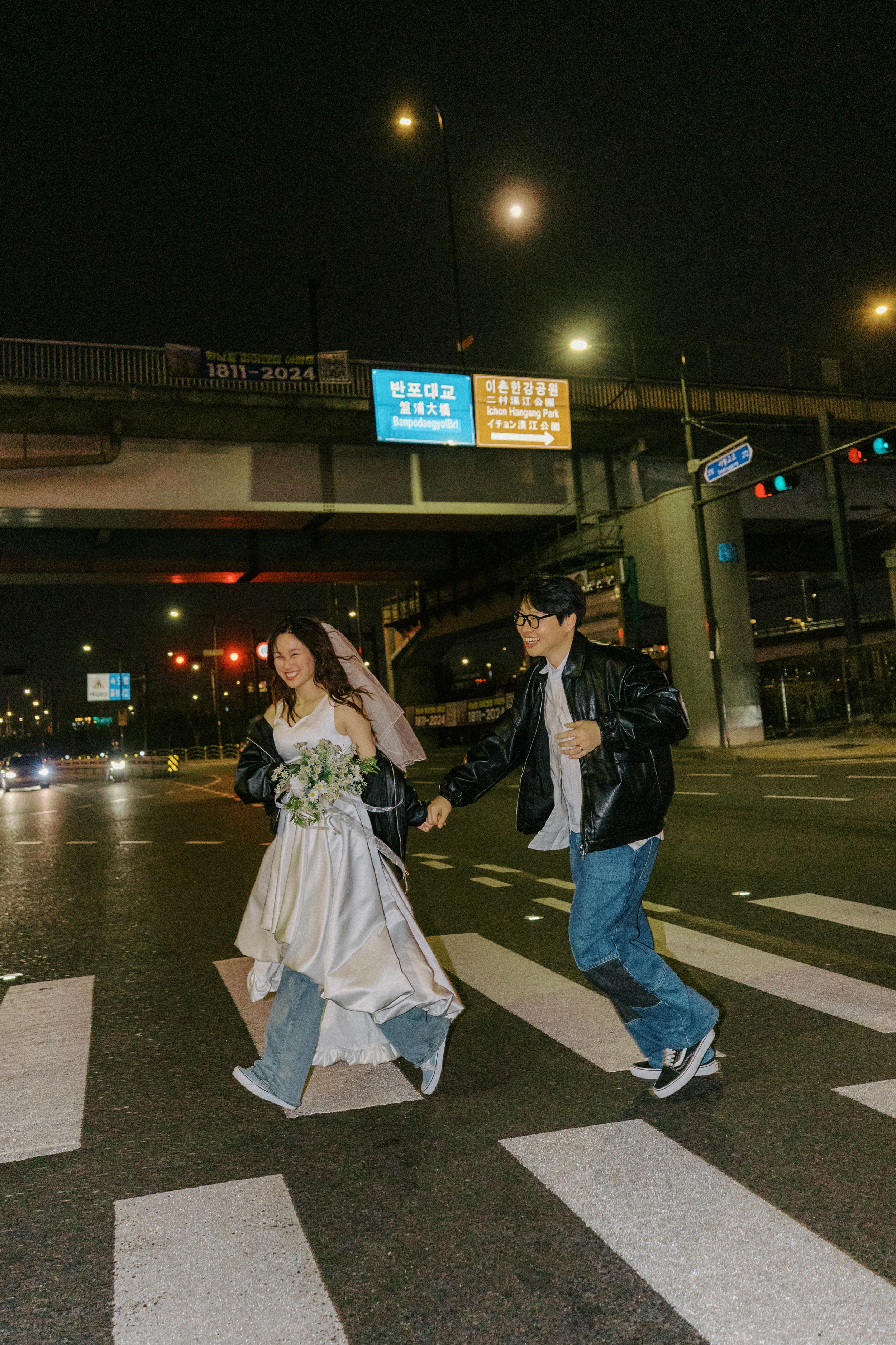 Couple crossing a city street at night, bride in white gown with bouquet, groom in leather jacket and jeans.