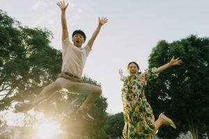 Two energetic figures leap joyfully in a sun-dappled garden with trees in the background.