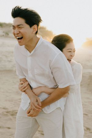 A joyful couple wearing white clothes laughs together on a sandy beach during sunset.