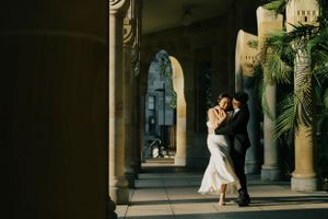 A couple in wedding attire dances romantically in an elegant columned archway with warm sunlight filtering through.