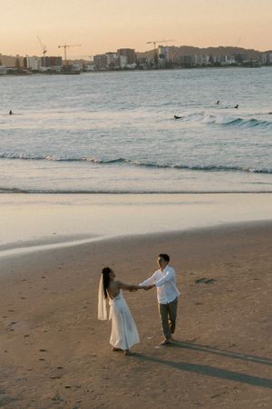 Two figures interact on a sandy beach at sunset with city buildings visible in the background.