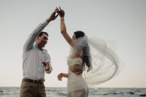 A couple dances together on a beach during sunset as the bride's veil flows gracefully in the wind.